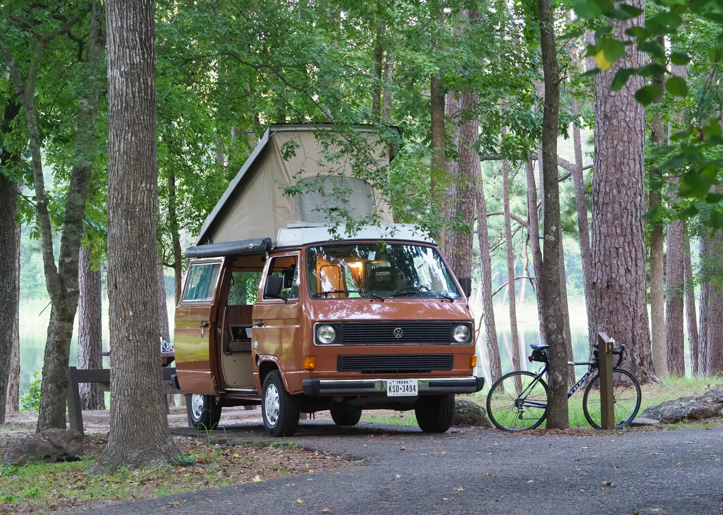 A brown VW camper van with pop-top extended, ideal for an RV rental, is parked in a scenic lakeside campground with a bike.