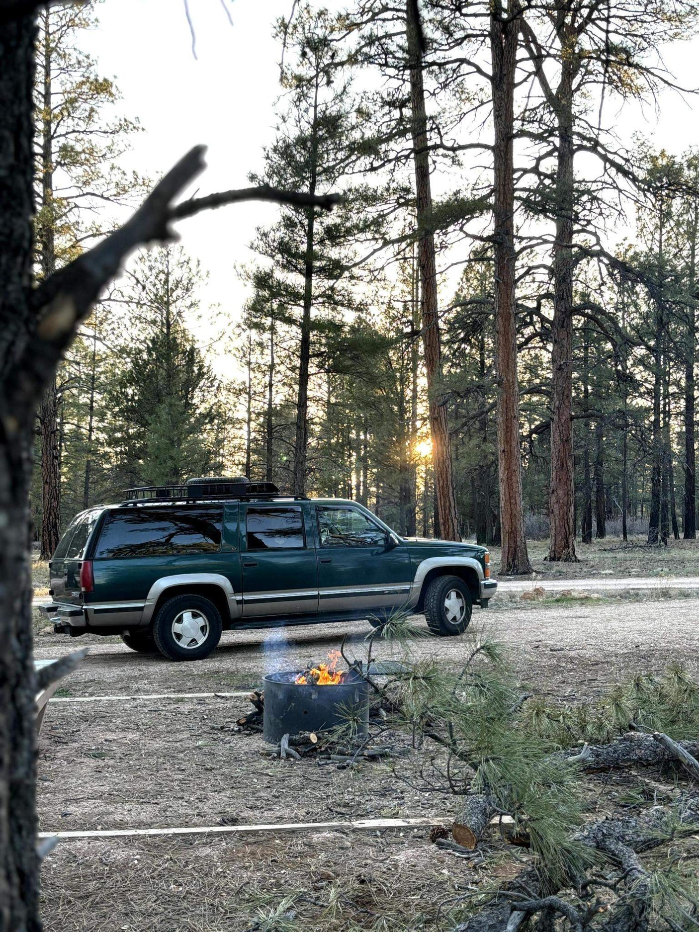 A dark green SUV is parked in a sunset-lit pine forest, a campfire burning warmly. Perfect for your next RV rental adventure!