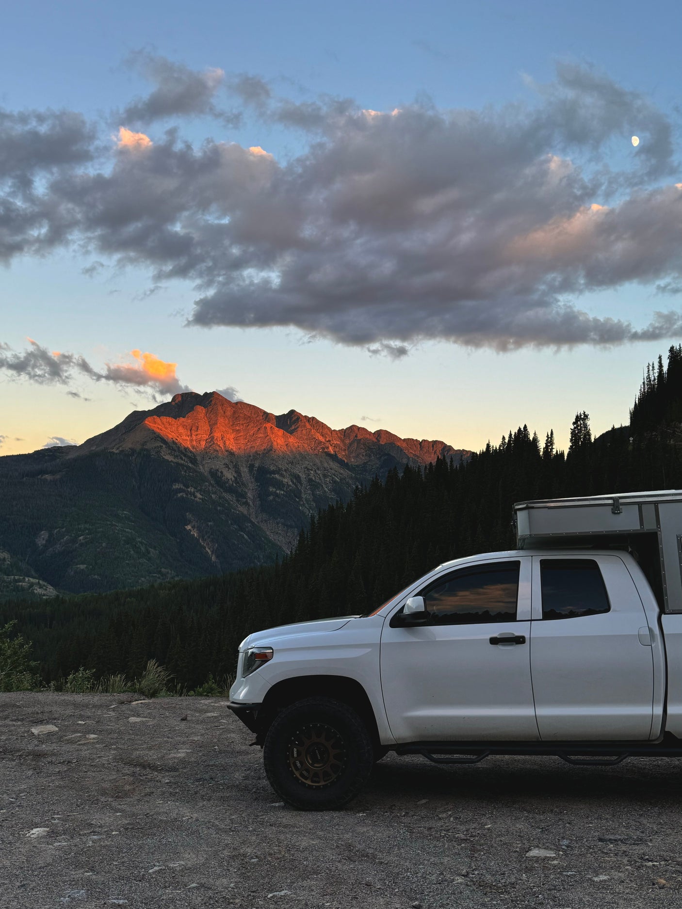 Enjoy a scenic RV rental trip! White truck camper at sunset with a moonlit mountain and forest backdrop.