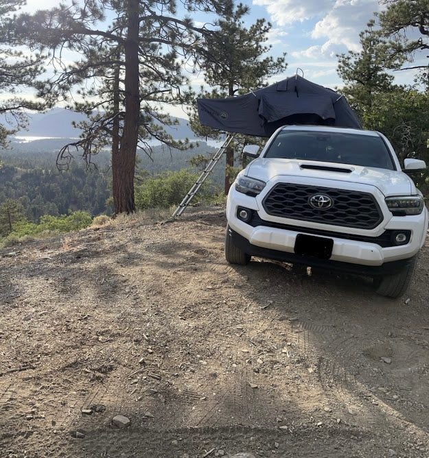 White Toyota Tacoma with rooftop tent set up on a dirt path, overlooking a serene lake. An adventurous RV rental option!