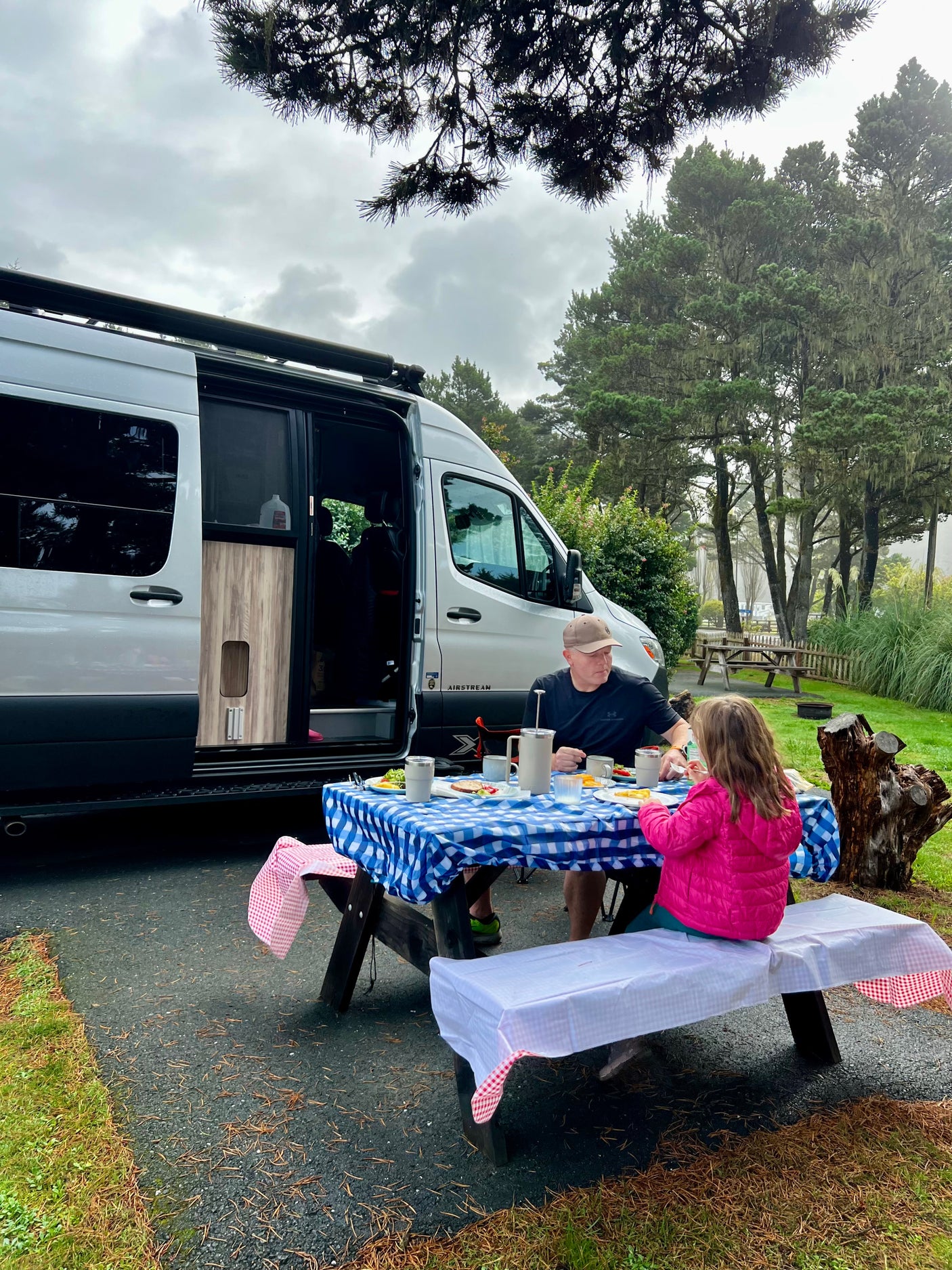 An RV rental trip: A man and child enjoy breakfast at a picnic table with a blue checkered tablecloth, next to a white camper van.