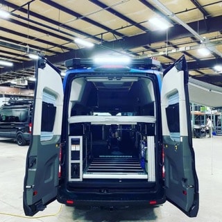 Inside a blue campervan, the rear doors open to reveal modular storage, a bed platform, and floor tracks, ready for RV rental adventures.