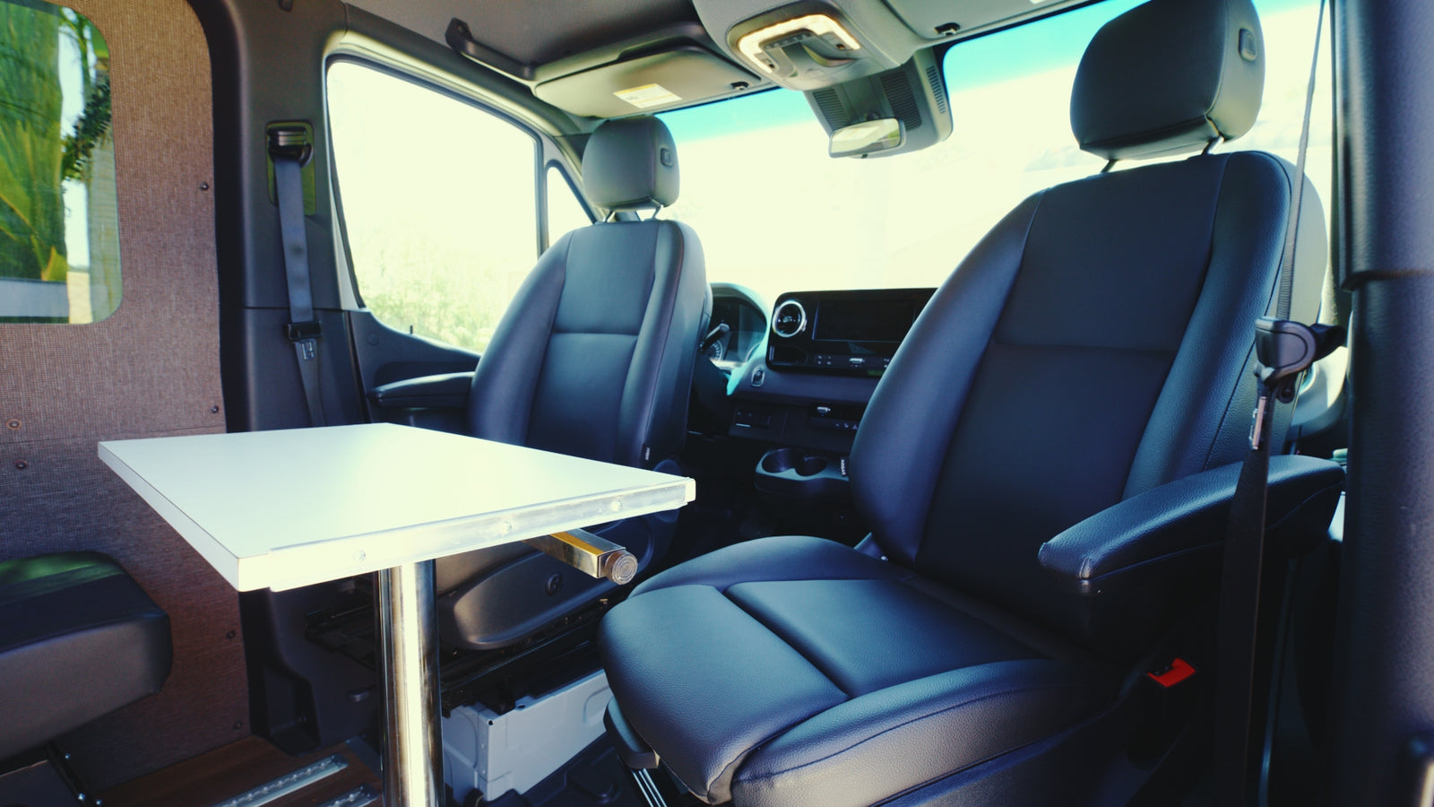 RV rental interior: two black front seats, a white table with chrome leg, and dashboard details visible, shot from the rear cabin.