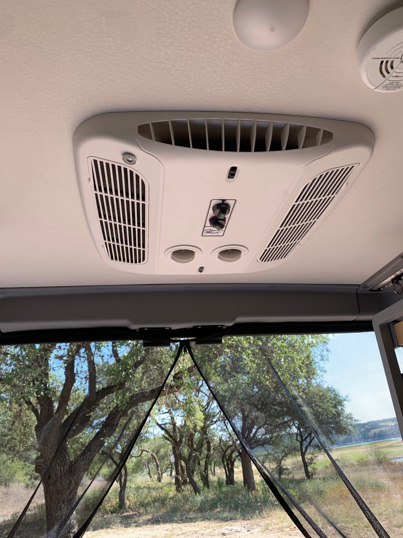 RV rental interior showing AC, smoke detector, and a view through mesh screen of trees & a lake.