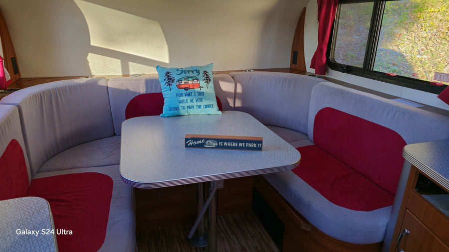 Cozy RV rental dinette: U-shaped seating in grey & red, with a humorous "Sorry" pillow & "Home" sign on the table.