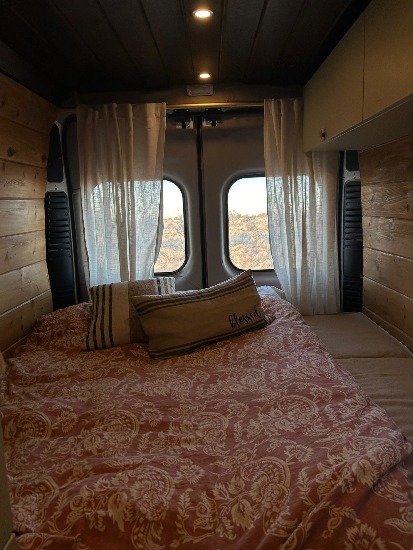 Cozy RV rental bedroom with a red patterned bed, wooden walls, warm lighting, and a scenic view of a dry landscape from the back windows.