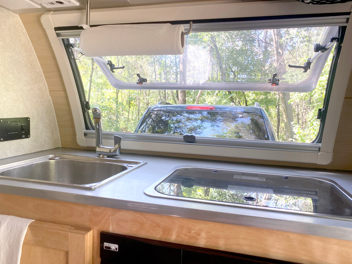 Interior of an RV rental kitchen with a sink, stovetop, and a large window overlooking a forest and a parked car.