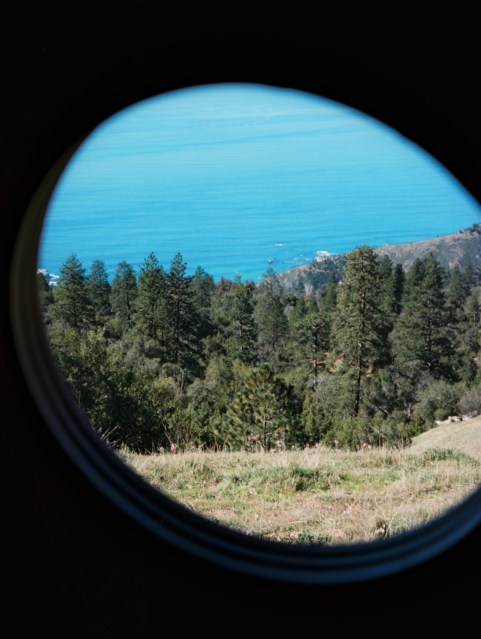 Stunning coastal view from an RV rental's round window: blue ocean, green mountains, and wildflowers on a grassy hill.