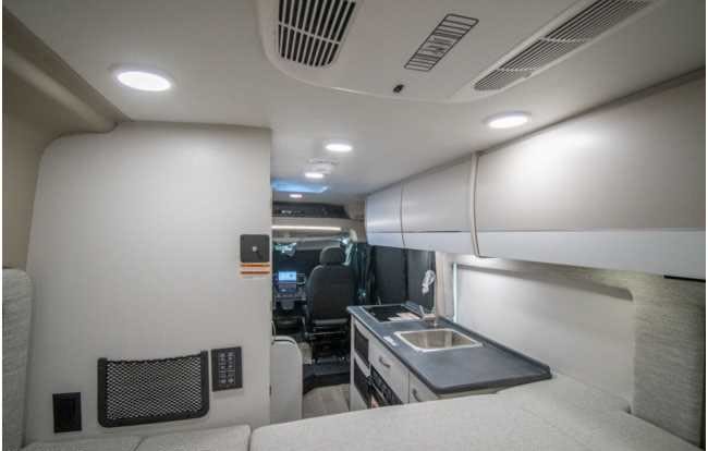 Modern rv rental interior with a compact kitchen, overhead storage, and a comfy bed, leading to the driver's cabin.