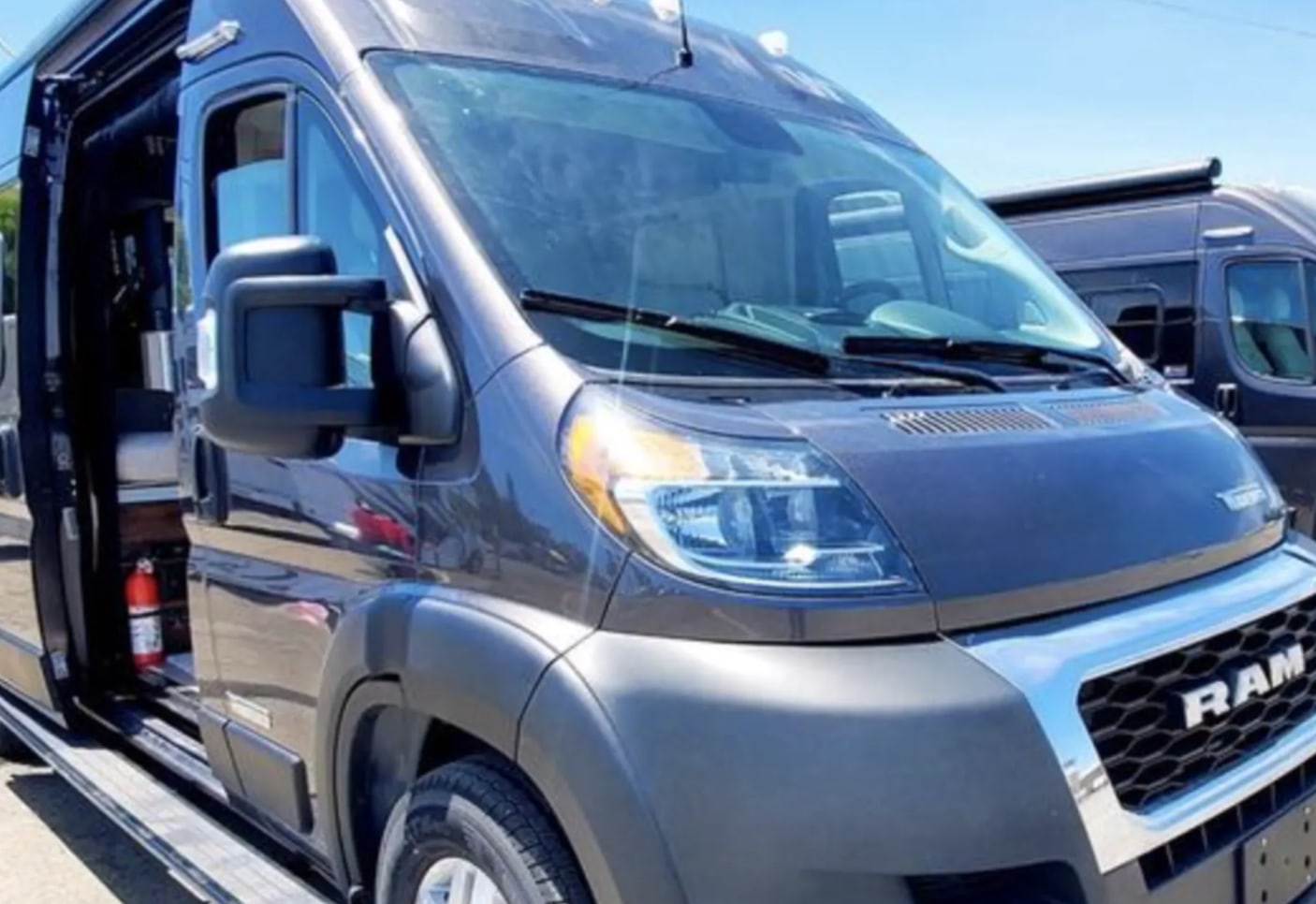 A gray Ram ProMaster RV rental, side door open to interior with fire extinguisher. Another van visible behind.