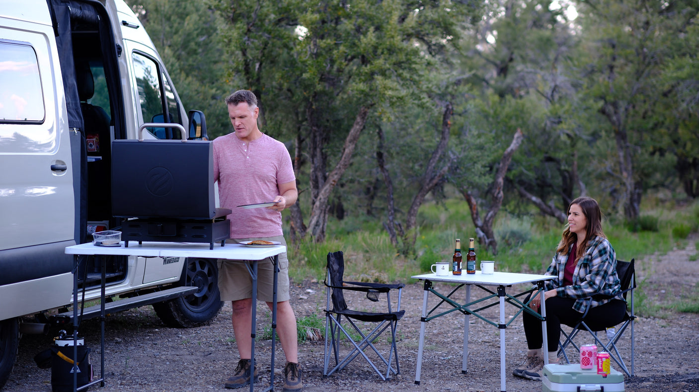 Couple camping with an RV rental: a man grills dinner next to their Winnebago van while a woman relaxes at an outdoor table.