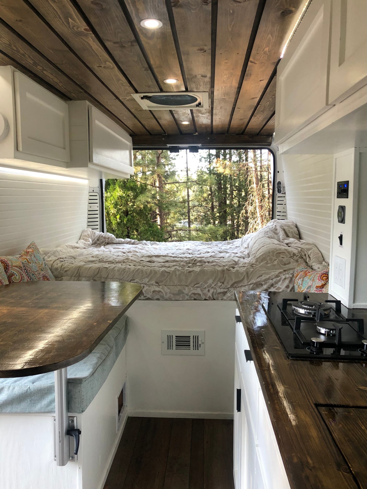 This RV rental interior features a cozy bed, kitchen, dining table, and a scenic view of nature.