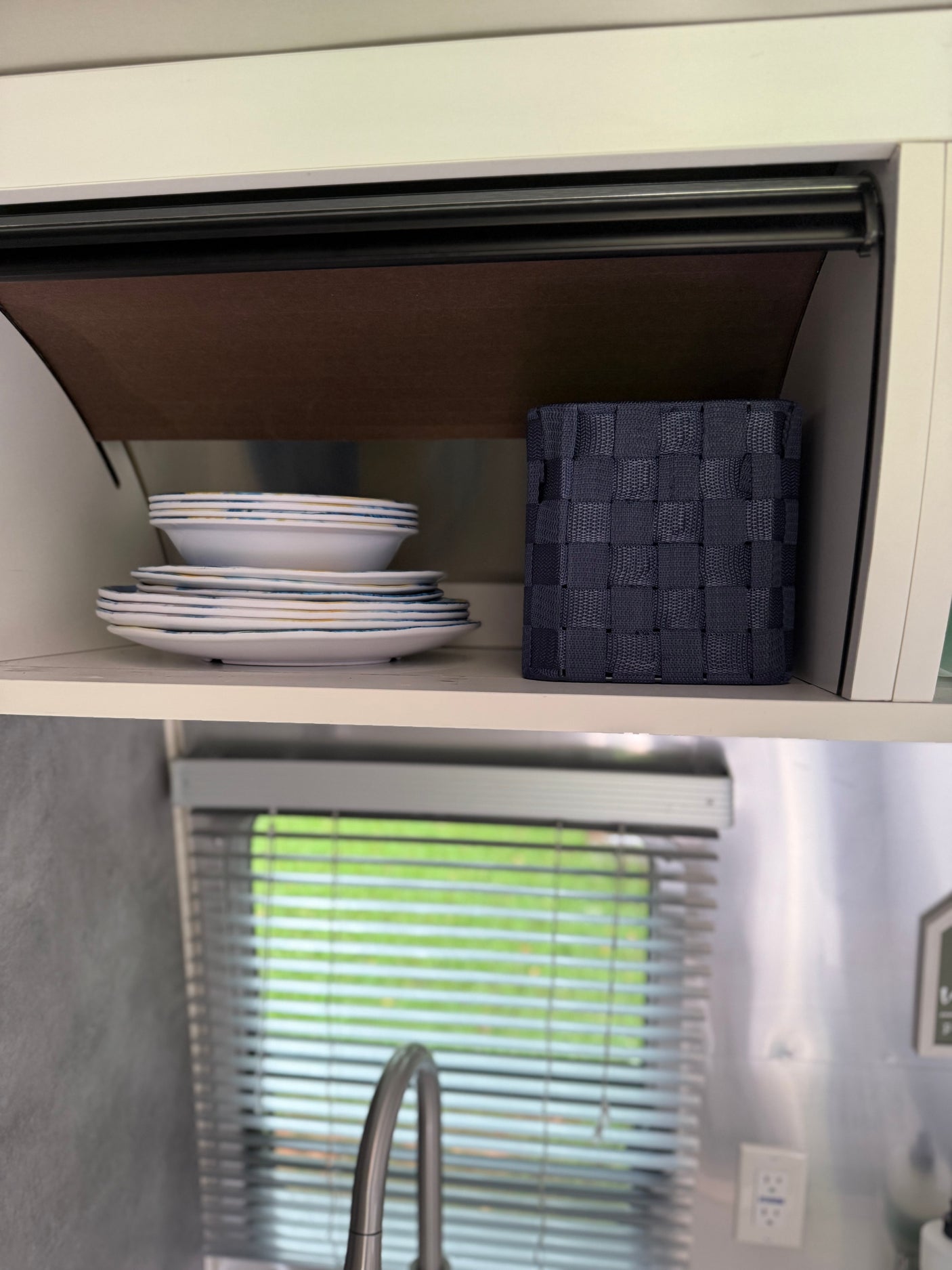 An RV rental's organized kitchen cabinet holds stacked dishes, a storage basket, and reveals a window with blinds and faucet below.