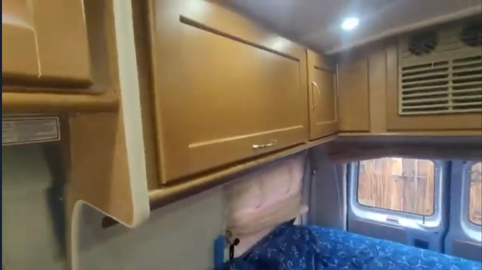 Inside an RV rental: light wood overhead cabinets, bright ceiling lights, a blue patterned bed below, and rear windows showing a fence.