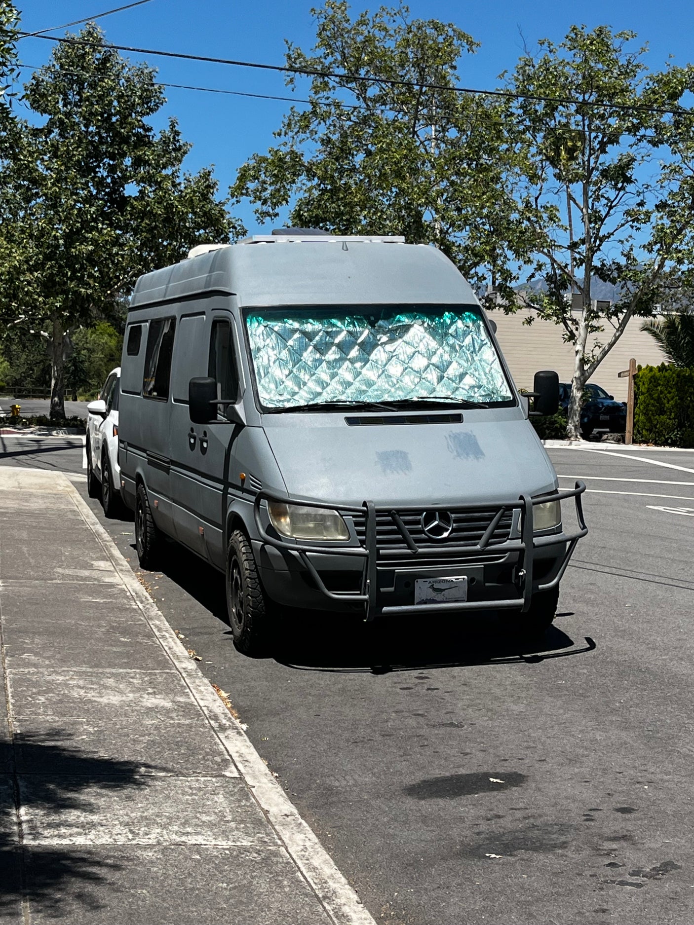 Grey Mercedes-Benz Sprinter camper van, possibly an RV rental, with a sunshade parked on a sunny street.
