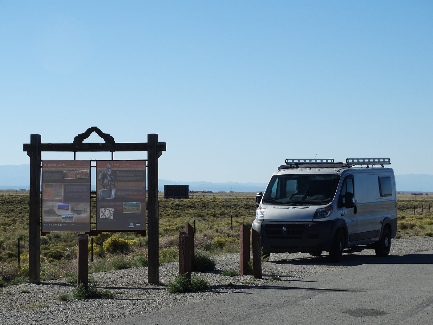 An RV rental camper van at a roadside historical sign ("On Sacred Ground") in a vast, sunny, open desert landscape.