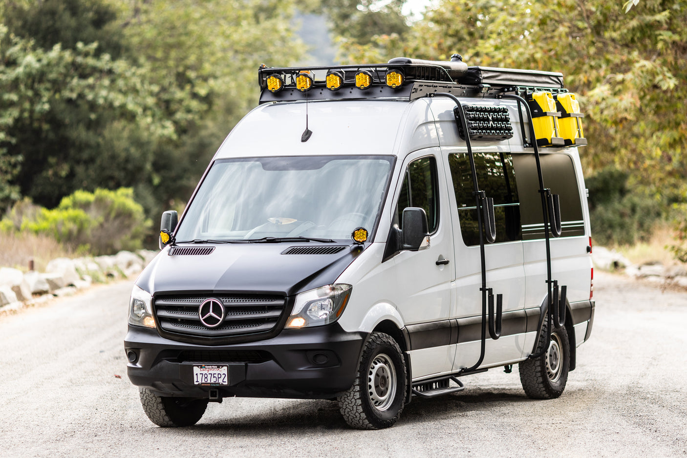 White Mercedes-Benz Sprinter adventure van RV rental, equipped with roof rack, lights, and off-road tires, parked on a road.