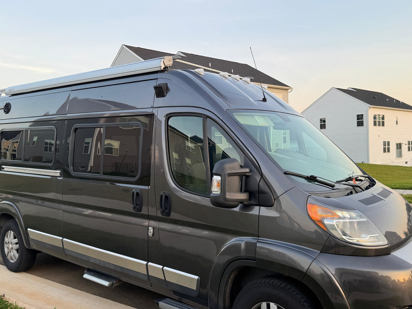 Gray camper van rv rental, ready for travel. Shows roof awning, side steps, and tinted windows in a residential setting.
