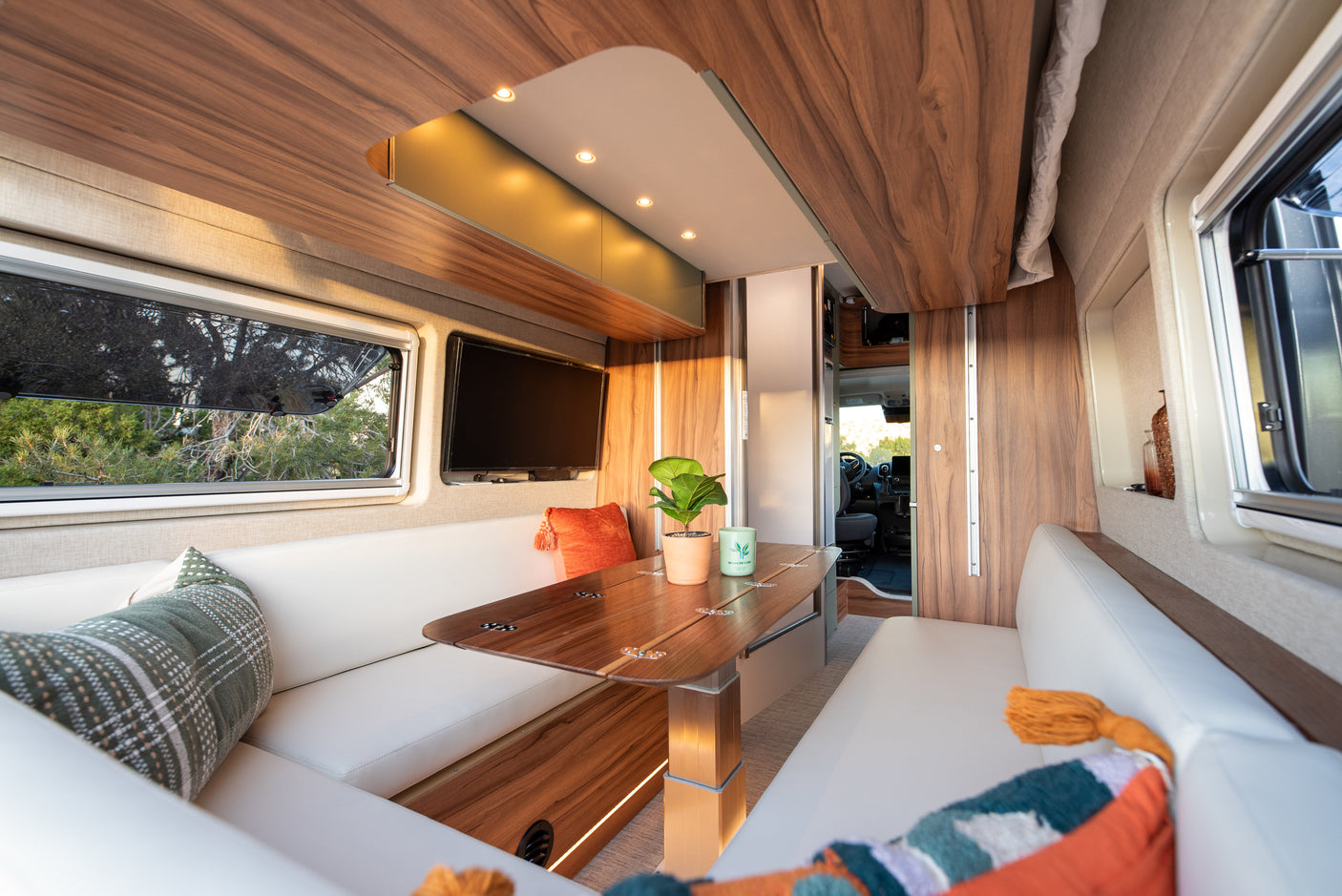 Cozy RV rental interior with a wooden dining table, white bench seating, TV, and vibrant pillows, offering a comfortable travel space.