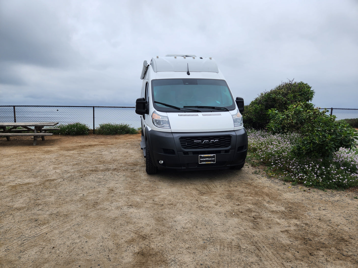 White camper van RV rental parked on a coastal dirt lot with ocean views, a fence, and picnic table.