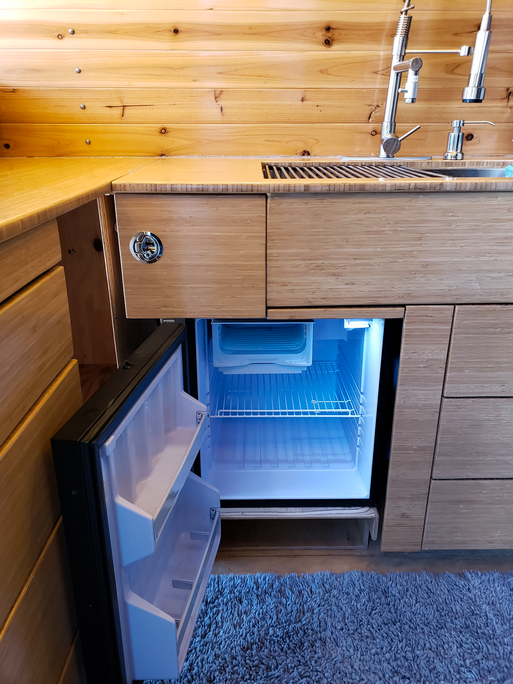 Modern RV rental kitchen featuring a compact fridge with a glowing blue interior, wood cabinetry, sink, and rug.