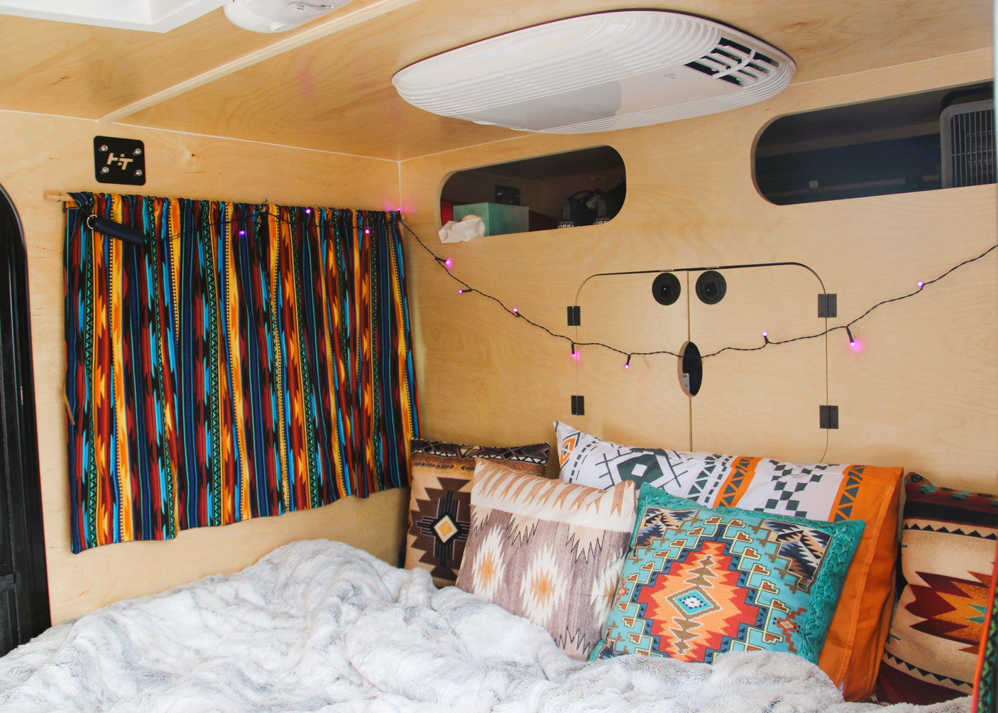 Cozy RV rental with a fluffy white bed, colorful Southwestern pillows, and purple string lights in a wood-paneled interior.