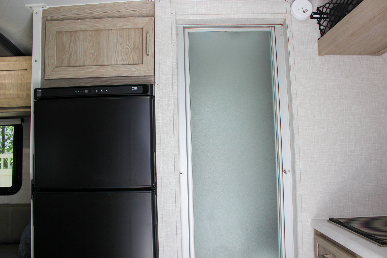 This rv rental interior features a black refrigerator, light wood cabinets, and a frosted glass shower door, ideal for mobile living.