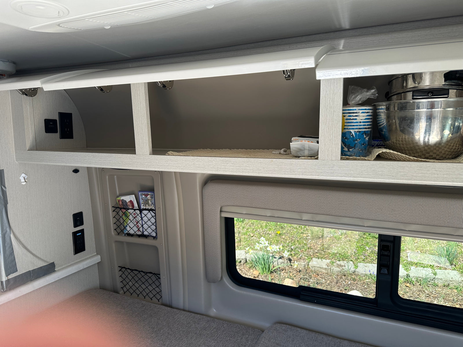 RV rental interior featuring open overhead cabinets with kitchenware, wall outlets, storage nets, and a window view to nature.