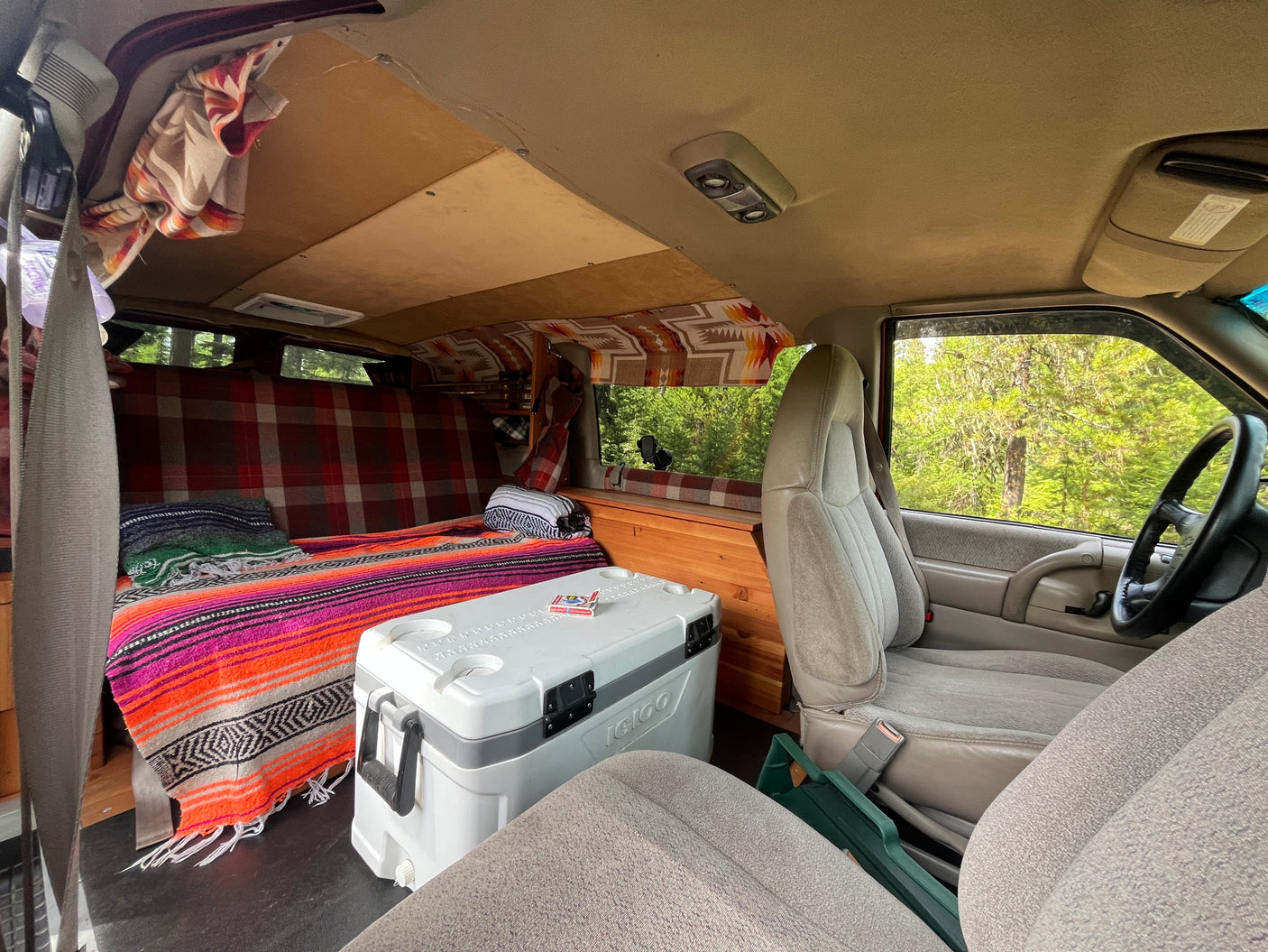 Cozy RV rental interior: Custom camper van features a built-in bed with colorful blankets, a cooler, and wooden storage.