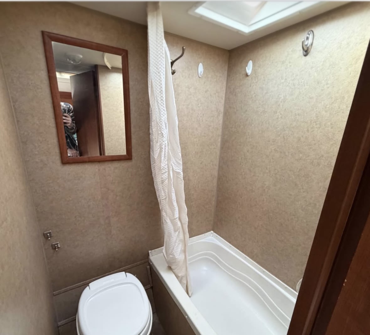 RV rental bathroom showing a compact toilet, shower, and tub combo with a mirror and hooks on textured walls.