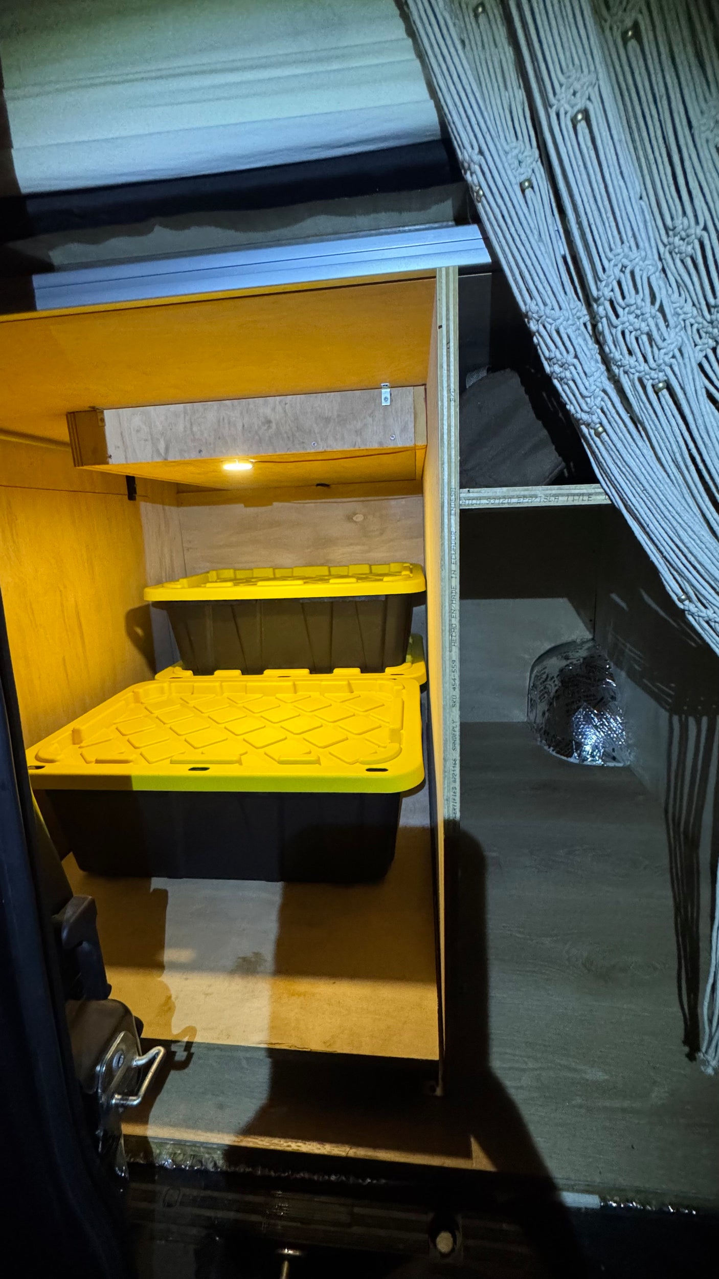 RV rental conversion: custom wooden storage unit with illuminated shelves, two yellow-lidded bins, and a macrame curtain.