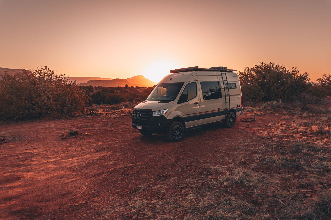 Scenic RV rental: a Sprinter van rests in a fiery desert at sunset, surrounded by low shrubs and distant mesas.