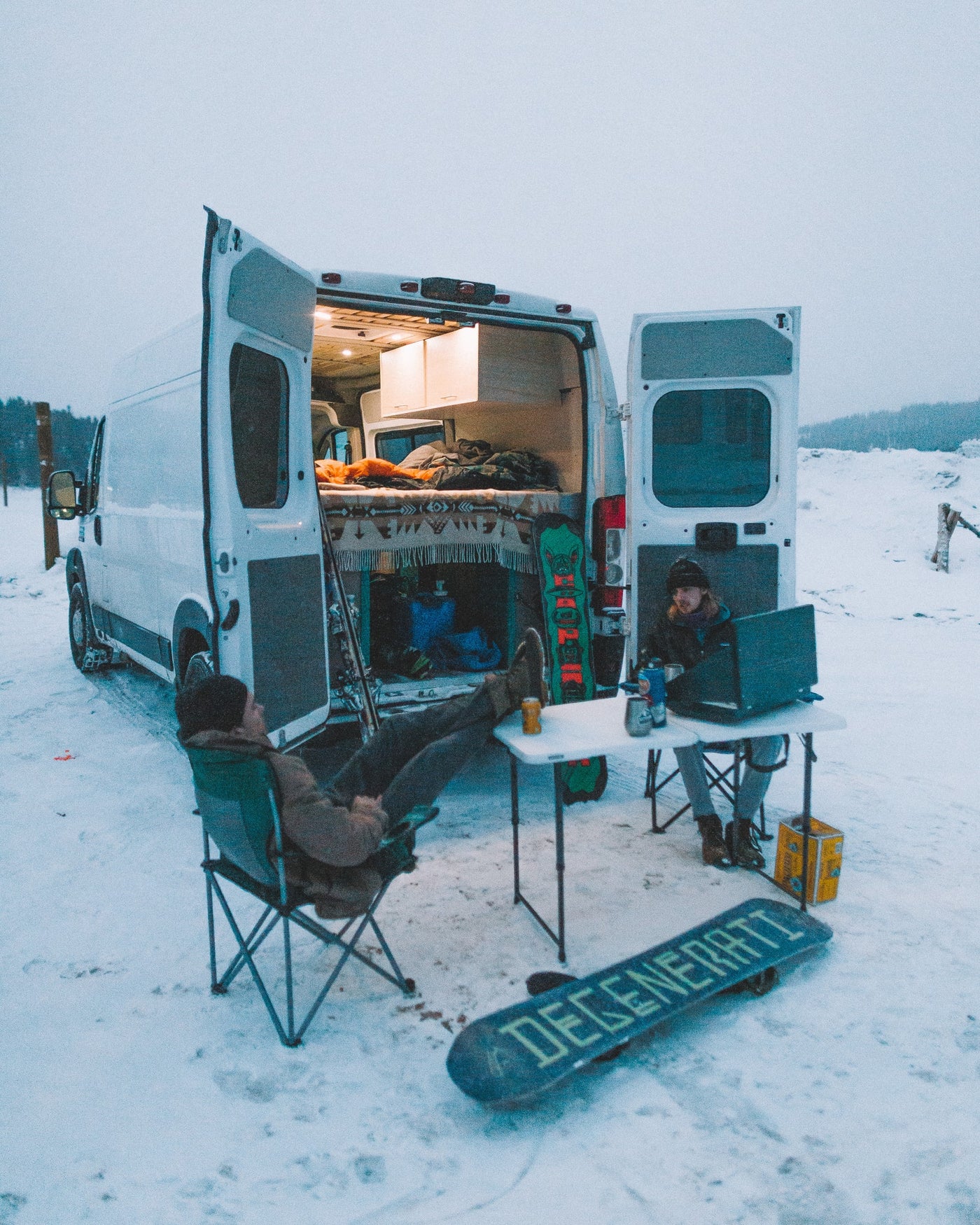 Cozy RV rental experience: Two people, one on a laptop, relax outside their camper van in a snowy landscape. Snowboards nearby. — photo 13
