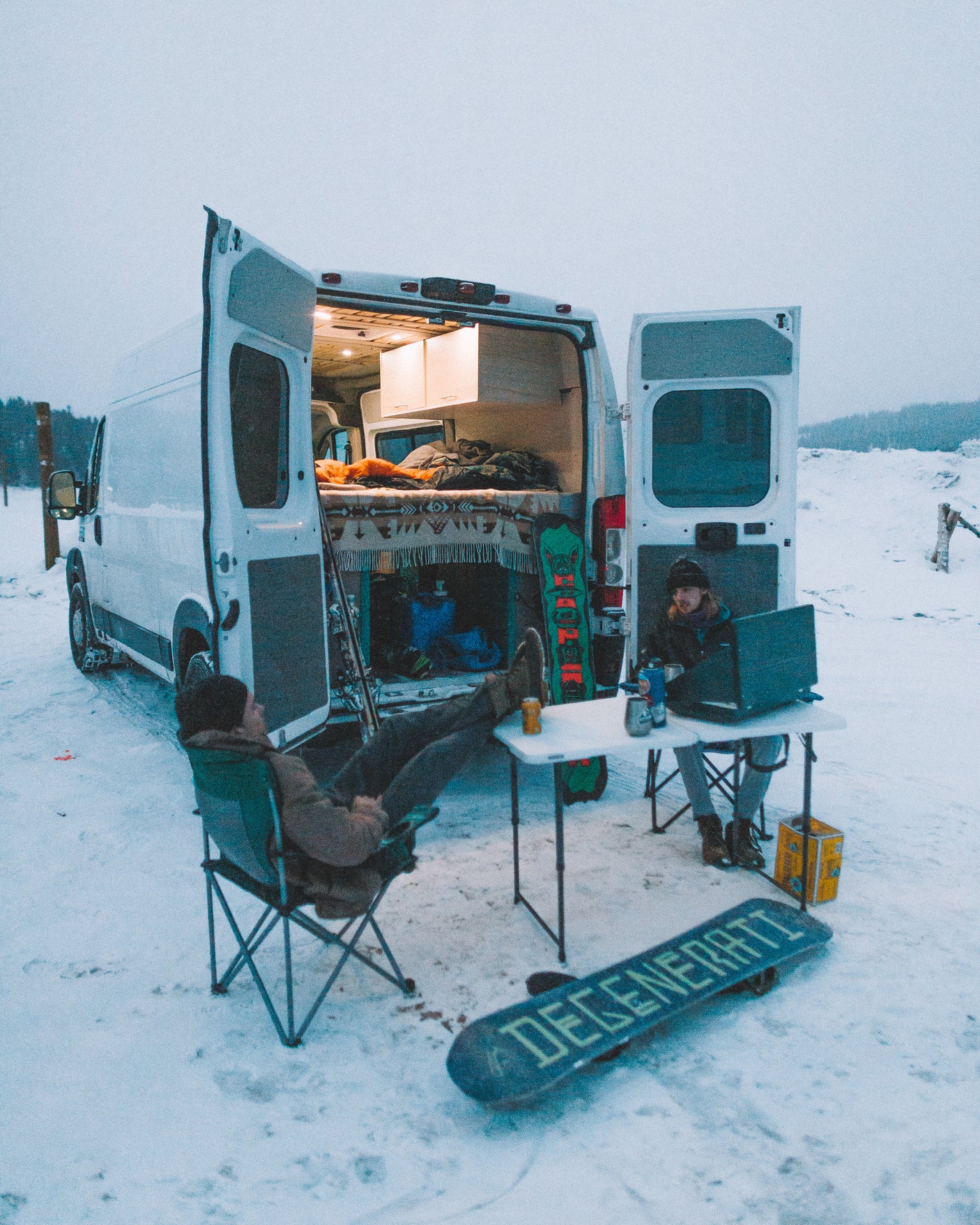 Cozy RV rental experience: Two people, one on a laptop, relax outside their camper van in a snowy landscape. Snowboards nearby.
