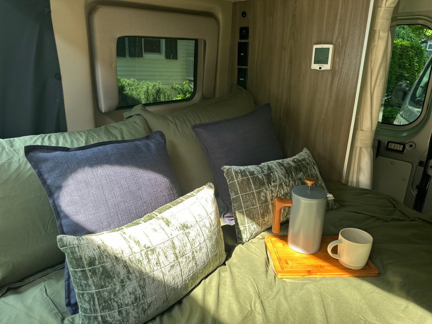 An RV rental's cozy bed with green sheets, blue & green pillows, and a coffee setup on a wooden tray by the window.