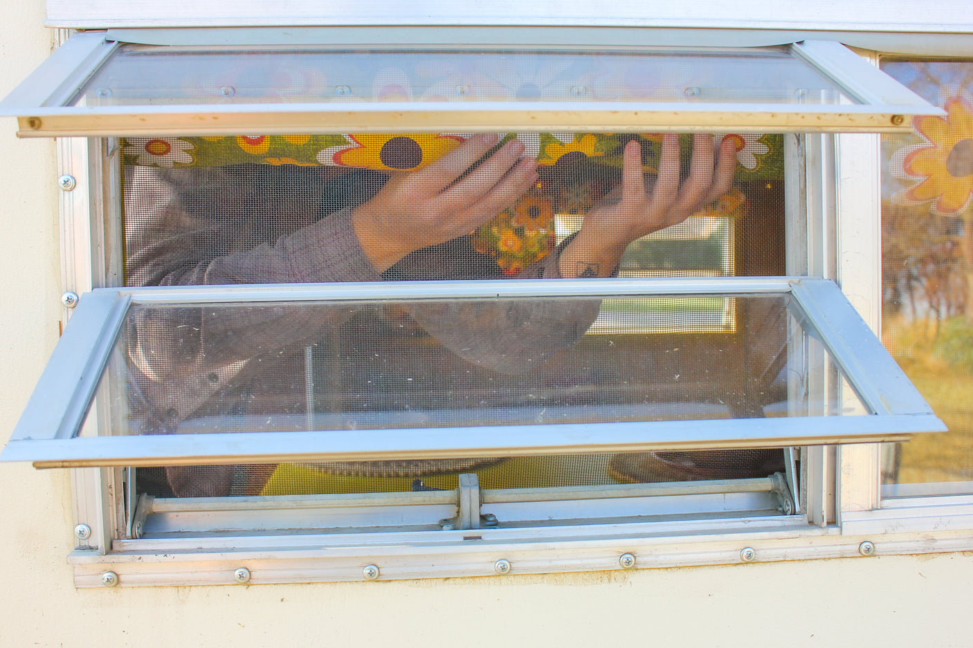 Through the screen of an RV rental window, hands are visible adjusting a colorful patterned item inside the vintage camper.