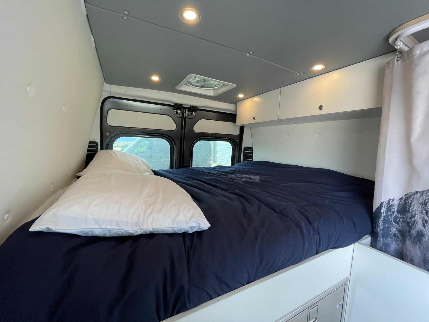 RV rental interior: A cozy bedroom with a dark blue bed, overhead storage, ambient lighting, and rear windows.