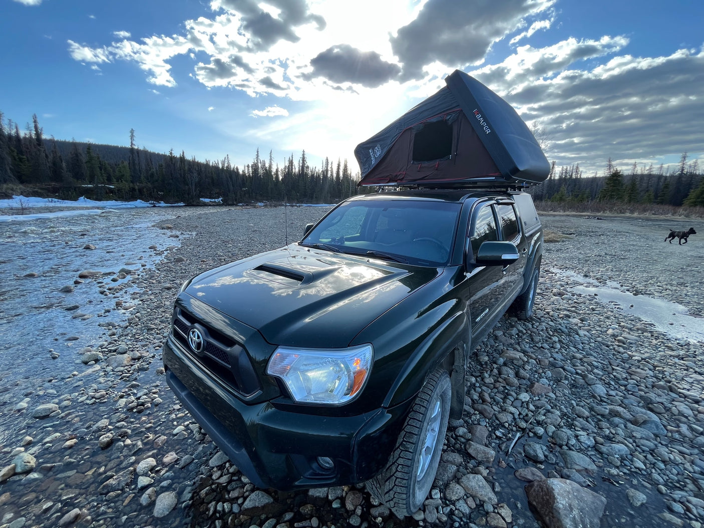 Adventure-ready dark green Toyota Tacoma with a rooftop tent on a rocky riverbank. Perfect for overland camping or an RV rental alternative.