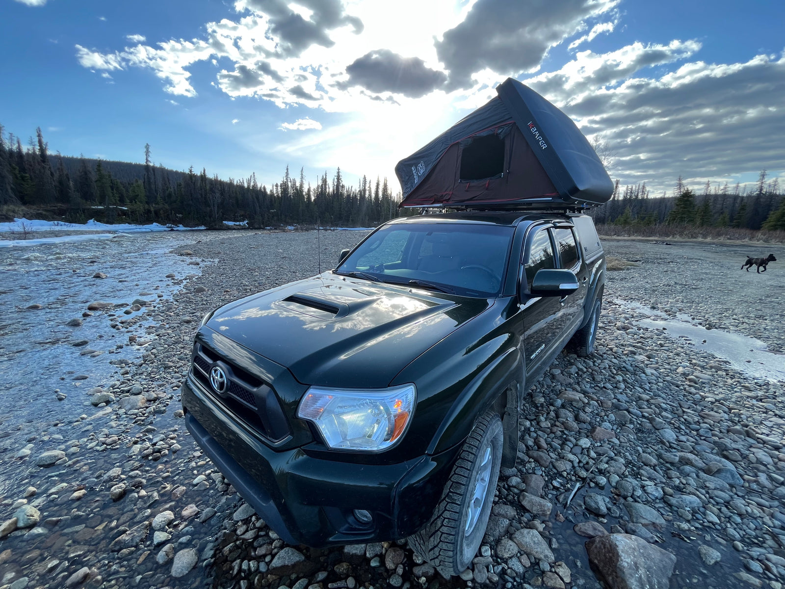 Adventure-ready dark green Toyota Tacoma with a rooftop tent on a rocky riverbank. Perfect for overland camping or an RV rental alternative.