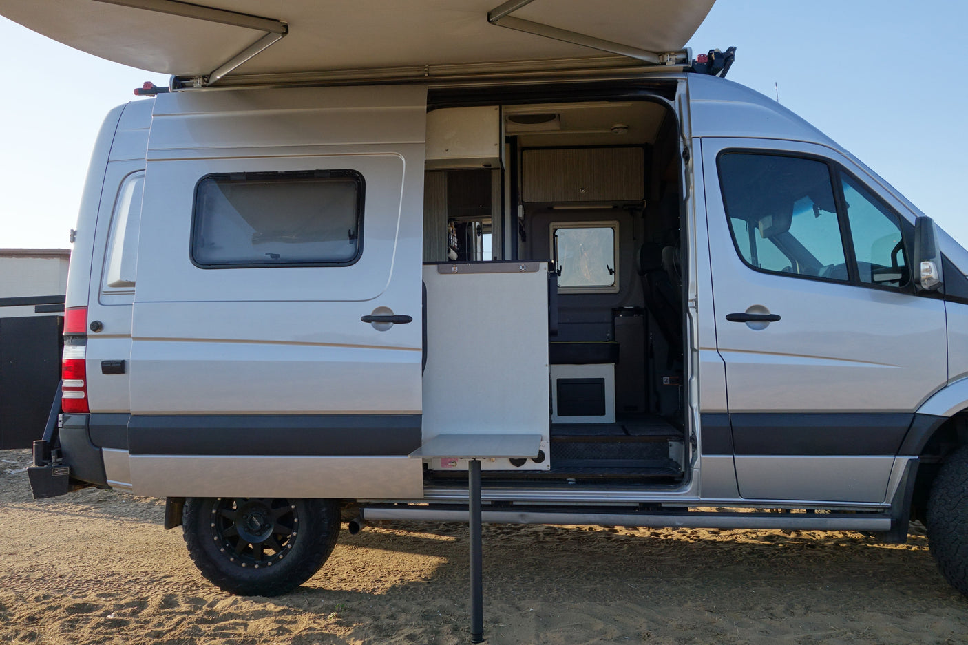 Silver camper van with open side door, extended awning, and table setup, ready for an RV rental trip or van life adventure.