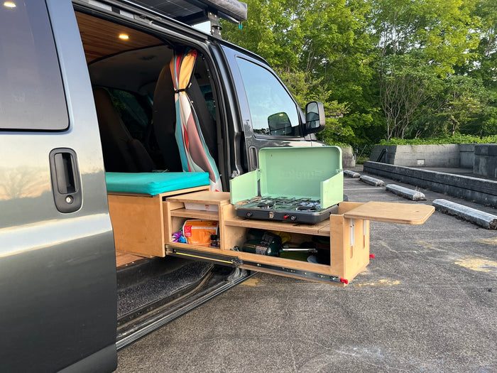 Open side door of an RV rental reveals a custom interior & a pull-out outdoor kitchen with a gas stove.