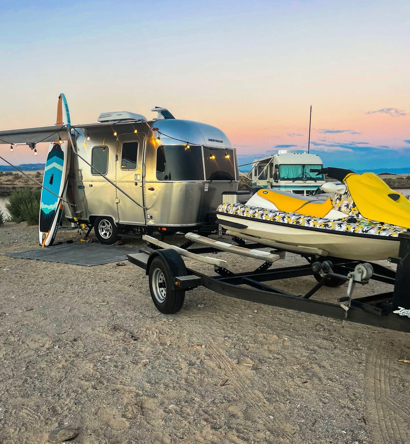 An Airstream RV rental, jet ski, and paddleboards are set up on sandy ground under a colorful sunset sky.