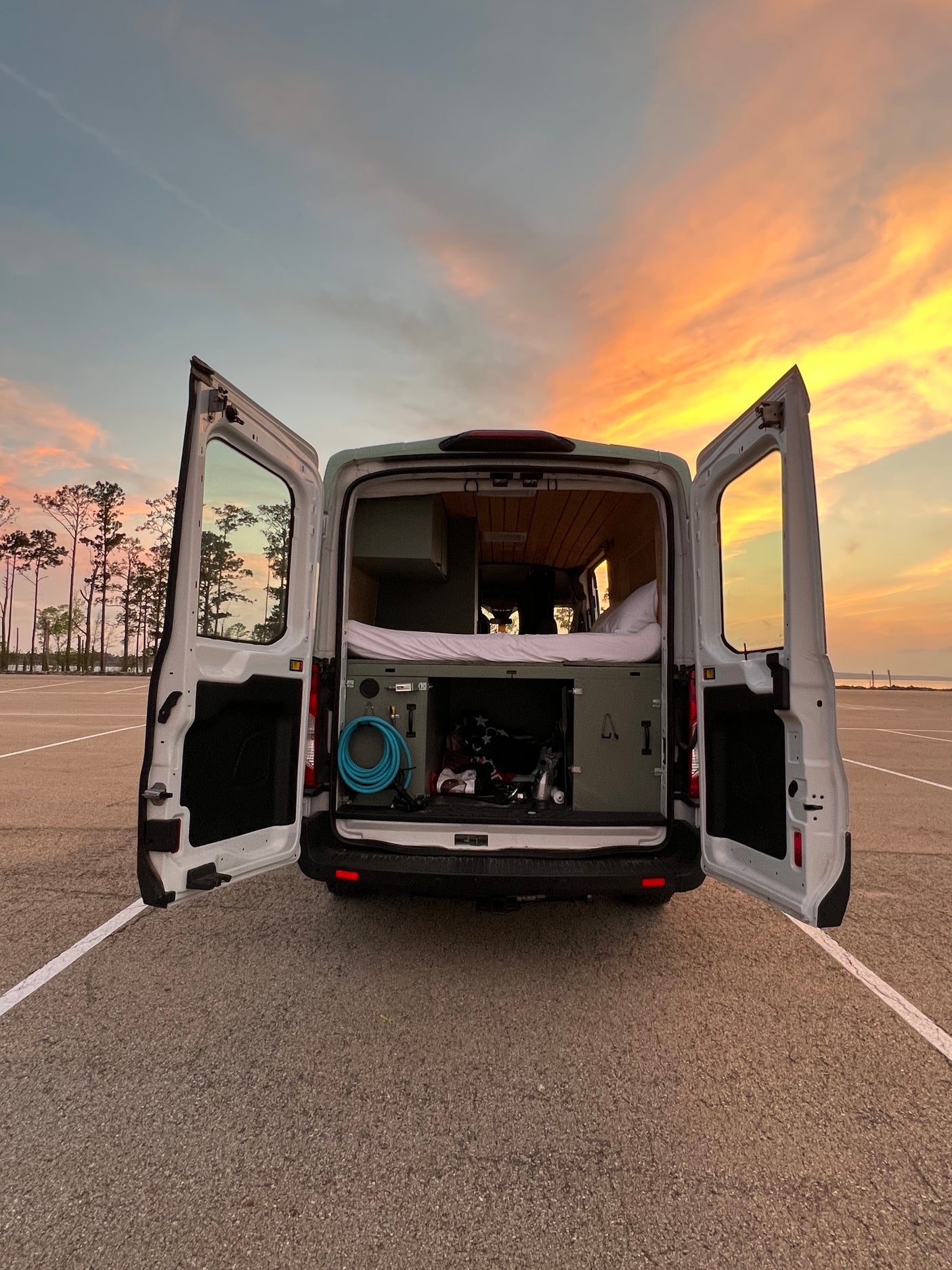 RV rental camper van's open rear reveals a cozy bed, interior storage, and gear. Golden sunset in the background.