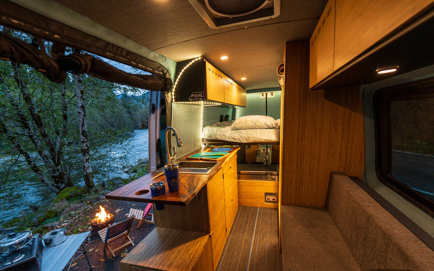 Cozy RV rental interior with kitchen, bed, and open door showing a serene river view and campfire. Ideal for nature escapes.
