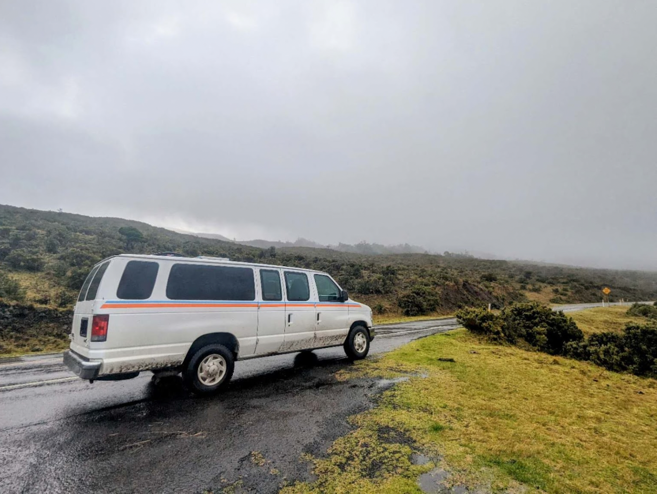 White RV rental van on a wet road, surrounded by misty hills and green scrubland. Perfect for a nature road trip.