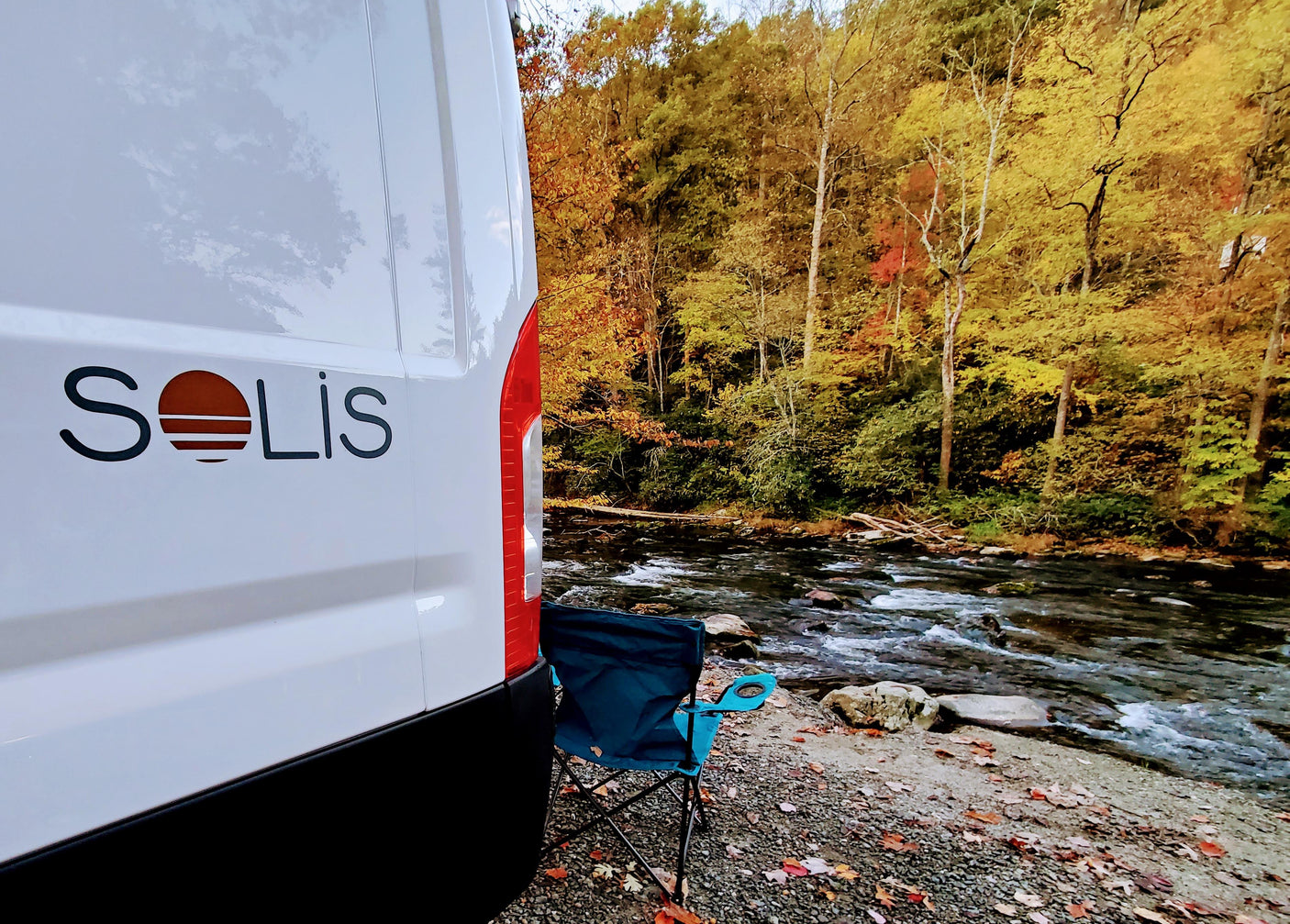 Solis RV rental by an autumn river. A camping chair offers views of colorful trees and flowing water.