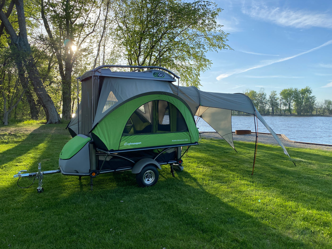 This compact SylvanSport trailer camper, ideal for an RV rental, is set up on lush green grass beside a scenic lake.