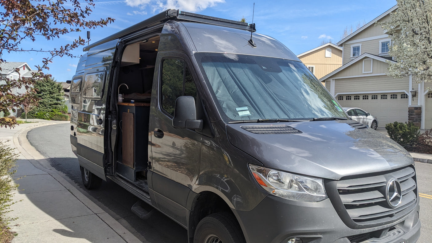 Custom dark gray Mercedes Sprinter camper van, an RV rental, parked on a residential street with its side door open revealing a kitchen.