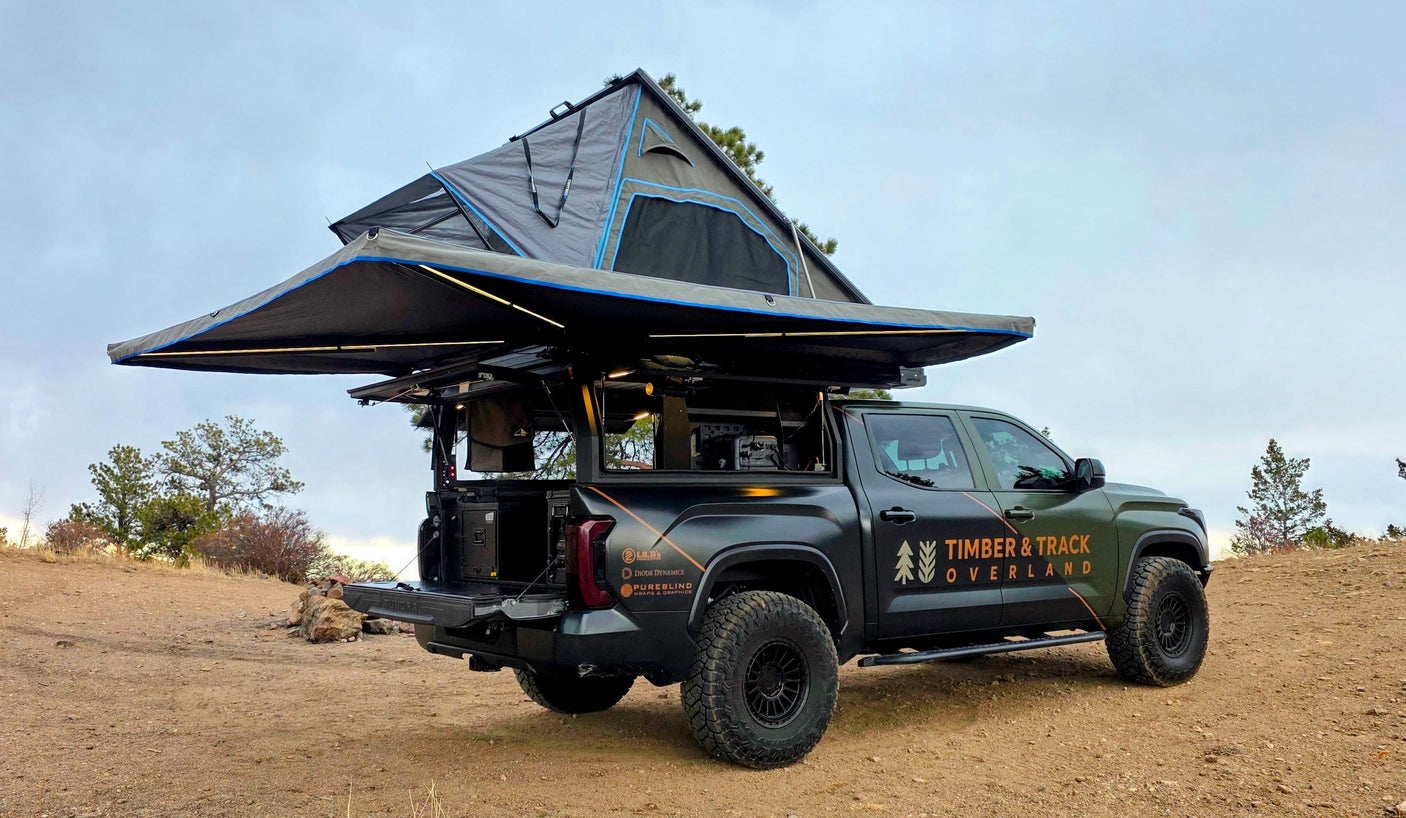 Dark green overland truck with deployed rooftop tent & awning on a dirt path. Ideal RV rental for off-grid camping adventures.