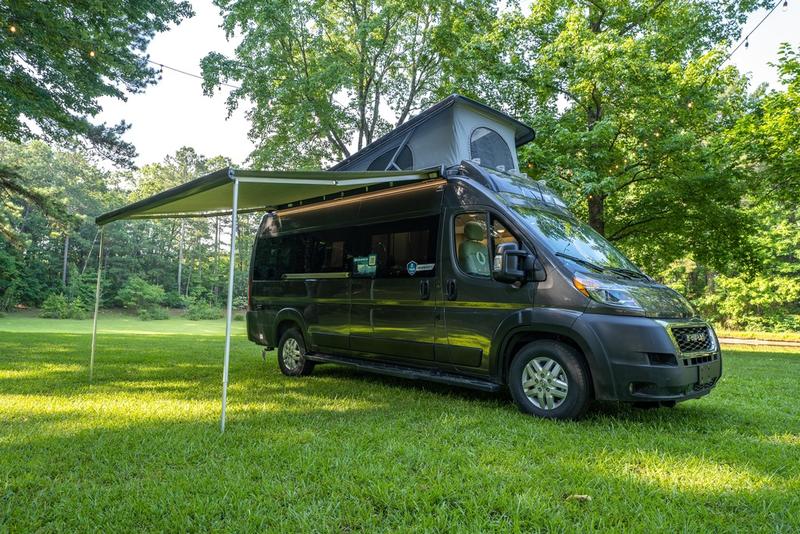 Explore in comfort! This dark gray RV rental camper van with pop-top and awning is ready for your next adventure in nature.|Ready for your RV rental adventure? Interior view of a comfortable camper van's front cabin and open outdoor scene.|Explore this RV rental's versatile camper van interior featuring a comfortable bench seat/bed